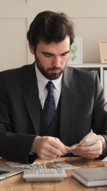 Bir ofiste çalışan ve nakit sayan, iş yatırımlarını ve kârları yöneten, profesyonel bir alanda refahı sağlayan odaklı bir işadamı. Dikey video.