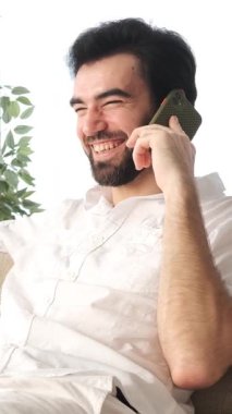 Evdeki bir telefon görüşmesi sırasında gülen mutlu bir adam, telekomünikasyon ve boş vakitlerle meşgul, sıcak bir ortamda rahatlama ve bağlantının tadını çıkarıyor. Dikey video.