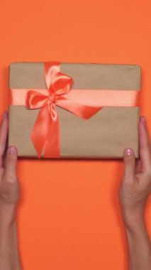 Elinde turuncu arka planda bir hediye kutusu olan bir kadın sürpriz bir paket sunuyor. Kutlama ve özel bir durum için hazırlanmış. Dikey video.