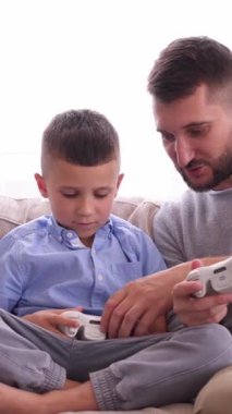 Mutlu baba ve oğul evlerinde gamepad kullanarak oyun oynuyorlar, eğlenceli ve eğlenceli bir ortamda birlikte eğleniyorlar. Dikey video.