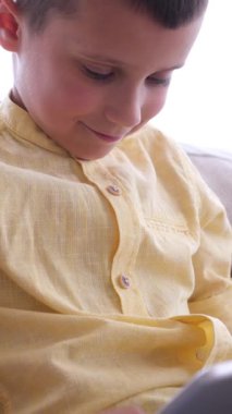 Neşeli bir çocuk evde online eğlence için tablet kullanıyor, rahat bir ortamda dijital aktiviteler yoluyla keyif ve rahatlama sağlıyor. Dikey video.