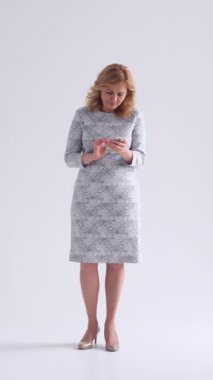 Beyaz arka planda cep telefonu kullanan nazik bir kadın, mesajlaşma ve sosyal medyaya bağlanıyor, rahatlama ve dijital bağlantı yayıyor. Dikey video.