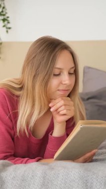 Memnun kadın evde bir yatakta dinlenirken kitap okuyor, boş zamanlarında öğrenme ve zevk gösteriyor. Dikey video.