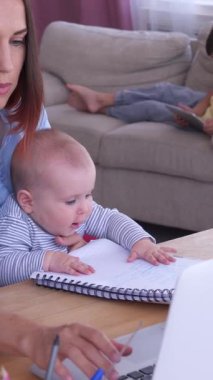 Evde çalışan ya da bilgisayarla e-öğrenim gören, kucağında bebek tutan, çevrimiçi eğitim peşinde koşarken anneliği kucaklayan pozitif bir anne. Dikey video.