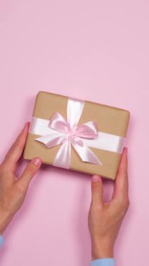 Pembe arka planda kurdeleli bir hediye kutusu hazırlayan bir kadın, özen ve detaylara dikkat ederek bir tebrik ya da selamlama hazırlıyor. Dikey video.