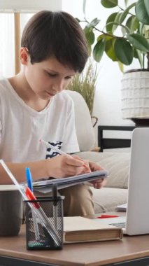 Düşünceli çocuk evde ödev olarak dizüstü bilgisayar ve not defteri kullanıyor, sessiz bir ortamda düşünceli bir şekilde eğitim ve öğrenim görüyor. Dikey video.