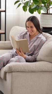 Evde oyalanmak için kitap okuyan tatmin olmuş bir kadın, sakin ve rahat bir ortamda keyif ve haz buluyor ve sakin ve meşgul bir tavır takınıyor. Dikey video.