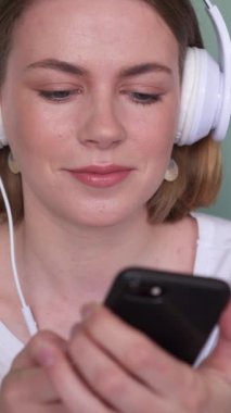 İnternette telefonla mesajlaşan, evde müzik dinleyen, gevşemiş bir ortam ile neşeli bir dijital etkileşime giren gülümseyen bir kadın. Dikey video.