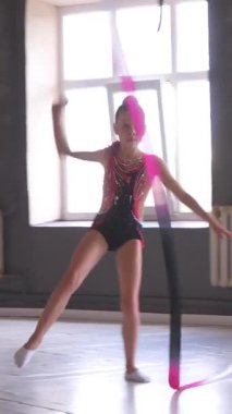 Sahne kostümlü bir kız stüdyoda kurdeleyle dans ediyor, sanatsal dans ifadesiyle performans odaklı bir ortamda şehvet ve jimnastik yeteneği sergiliyor. Dikey video.