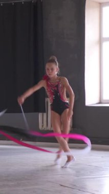 Sahne kostümü giymiş bir kız stüdyoda kurdeleyle dans ediyor, dans performansına ve öğrenmeye odaklanıyor, koreografi odaklı bir ortamda sanatsal yeteneğini sergiliyor. Dikey video.