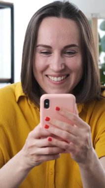 Mutlu bir kadın çevrimiçi eğlence için telefon kullanıyor, evde sosyal medyada sohbet ediyor, rahat bir dijital ortamda neşeli bir iletişim kuruyor. Dikey video.