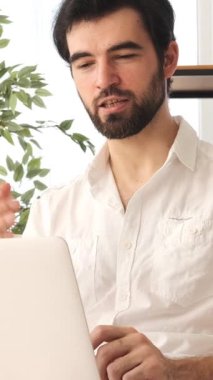 Dost canlısı bir adam uzaktan eğitim sırasında evde dizüstü bilgisayarla çevrimiçi bir video görüşmesi yapıyor ve içeride coşkuyla eğitim toplantılarına katılıyor. Dikey video.