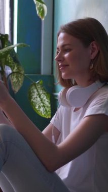 Gülümseyen bir kadın evde boş zamanlarında dinlenirken ve kulaklıkla müzik dinlerken sakin ve neşeli bir anın tadını çıkarıyor. Dikey video.