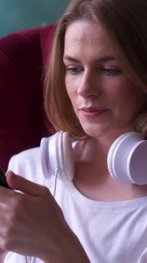 İnternette boş vakit geçirmek için cep telefonu kullanan ve internette gezinen pozitif bir kadın rahat ve neşeli bir dijital eğlenceye girişiyor. Dikey video.