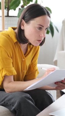 Uzaktan kumandalı bir işçi evdeki online finans işleri için dizüstü bilgisayar ve evrak işlerini kullanıyor, rahat bir çalışma alanında gayretli bir şekilde işlerle uğraşıyor. Dikey video.