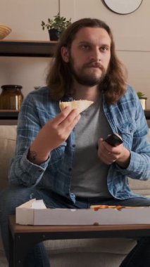 Akşam evde boş vakitlerinde pizza yiyip televizyon izleyen, rahat ve rahat bir atmosferde rahatlayan bir adam. Dikey video.