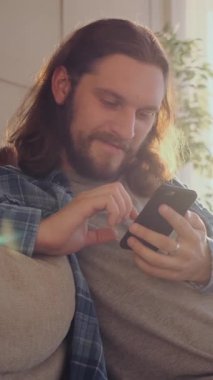 İnternet eğlencesine dalmış memnun bir adam, evde dinlenirken çevrimiçi eğlence için telefon kullanıyor, rahat ve ilgi çekici bir dijital deneyimin tadını çıkarıyor. Dikey video.