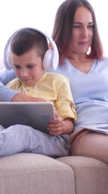 Sıcak ve iç açıcı bir atmosferde anne ve oğlu, evde online boş vakit geçirmek için tablet bilgisayar ve dizüstü bilgisayar kullanıyor, dinleniyor ve dijital aktivitelerin tadını çıkarıyor. Dikey video.