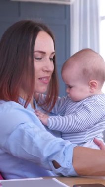 Gülümseyen anne kucağında bebekle evde dizüstü bilgisayarla çalışıyor, sıcak ve samimi bir ortamda internet e-öğrenimi yapıyor. Dikey video.