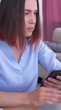 Evde dizüstü bilgisayar ve telefonla çalışan odaklanmış bir kadın, çocuklar arka planda oynarken işleri idare eden, iş ve aile hayatını dengeleyen bir kadın. Dikey video.