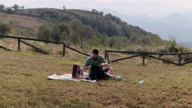 Bir baba ve genç kızın piknik battaniyesinde rahatlarken ve bir dağ çayırında çerezleri paylaşırken gerçek zamanlı bir fotoğraf..