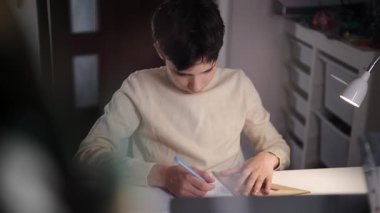 Ev masasında, bir lambanın altında grafik kağıdına geometrik çizgiler çizen bir gencin orta ön görüntüsü..