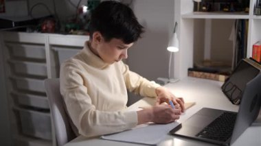 Genç bir çocuğun ev masasında oturup dizüstü bilgisayar ve masa lambasının yanında ödev yazışının yan görüntüsü..