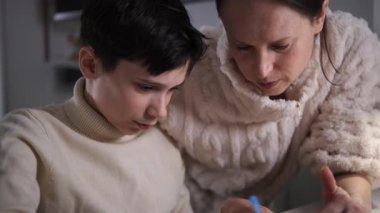 Bir annenin ergen oğluna ödevlerini evdeki bir masada deftere yazarken bir görevi açıklarken yakın plan görüntüsü..