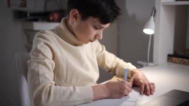 Masada oturan ve evdeki masa lambasının altındaki bir defterde ödev yazan genç bir çocuğun yan görüntüsü..