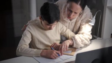 Evde bir masa lambasının altına geometri çizerken genç bir çocuğun üzerine eğilen bir annenin yan görüntüsü..