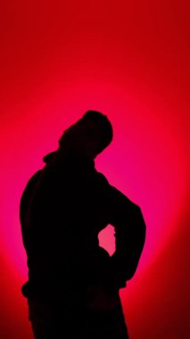 Kapüşonlu bir adam canlı kırmızı bir arka planda modern sokak dansı yapıyor. Minimalist bir stüdyoda temiz bir siluete aydınlatılıyor..