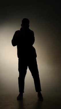 Kapüşonlu bir adam, minimalist bir stüdyoda temiz bir siluet olarak çekilen, sıcak bir arka plan gradyanı karşısında gerçek zamanlı olarak modern sokak dansı yapıyor..