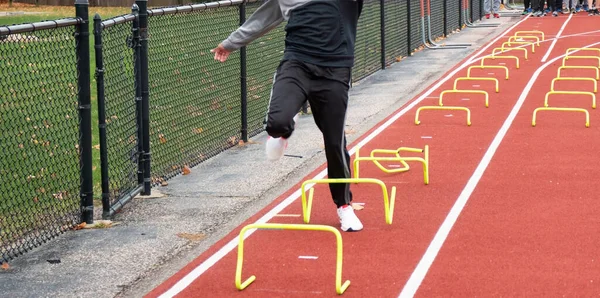 A high school boy is tripping over yellow mini hurdles while running a sports drill in lane on a red track at practice.