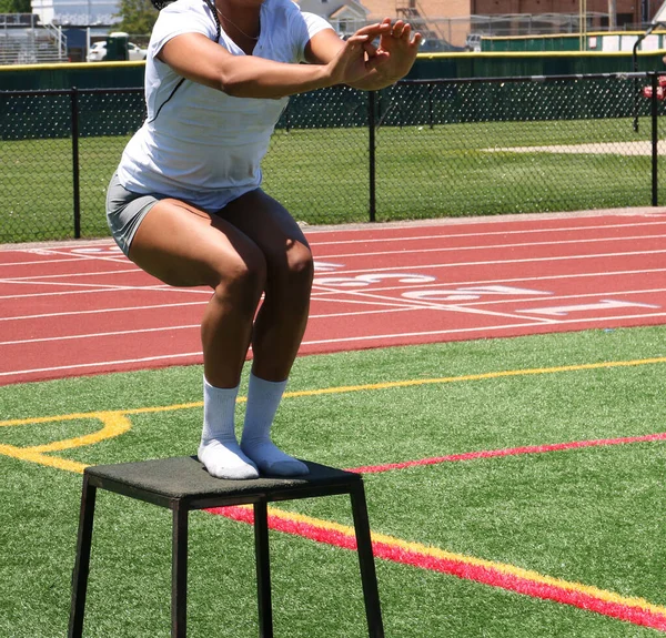 A female college athlete landing on to a plyos box with exercising on the field next to a track.