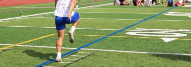 One football athlete running though a ladder drill while others rest on the ground in the background during summer preseason training camp.