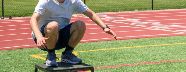 A male high school teenager landing on a ploys box in a squat position on a turf field at summer sports camp.