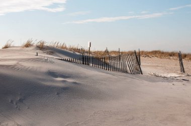 Side view of a sand dune with the sand smooth fron the wind and burying parts of the picket fence with a keep off the dubes sign in the background.