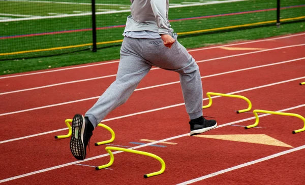 Side view of a high school boy running the wicket drill over yellow mini banana hurdles on a red track during track practice.