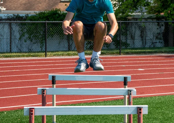 Teenage boy jumping over track hurdles for agility and strength during track and field practice in the summer.
