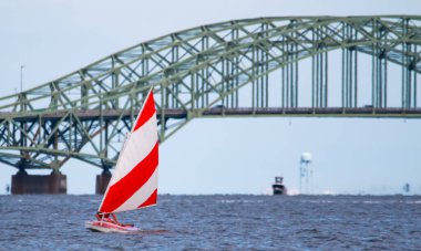 Front view of a close up of one red and white sunfish sailboat sailing on the Great South Bay with the bridge close in the background.
