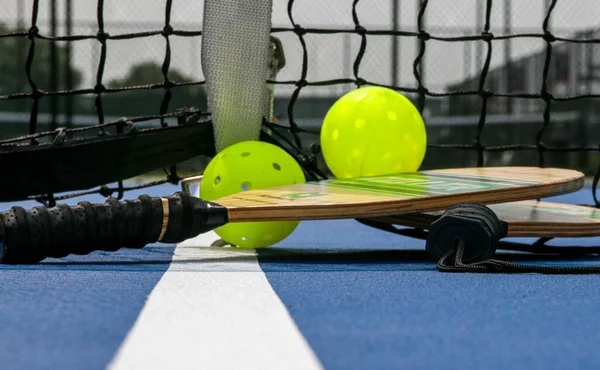 Close up of two pickleball rackets and two yellow whiffle balls next to the net on a tennis court.