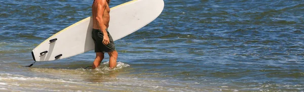 一个男冲浪手腋下夹着冲浪板走进大海在长岛冲浪的侧视图 — 图库照片