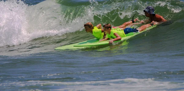 ギルゴビーチ ニューヨーク アメリカ 2023 サーフキャンプカウンセラー 彼らのサーフボードにネオングリーンシャツに2人の男の子を保持し ギルゴビーチのバンガーサーフキャンプ中にサーフィンに行く波をキャッチします — ストック写真