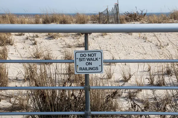 A sign that is on a metal fence soys do not sit or walk on railing in the dunes at a beach on the ocean