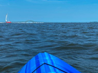 Uzakta bir köprüsü olan sakin, berrak bir mavi körfezde bir kişi kano kullanıyor. Gökyüzü parlak bir mavi ve güneş parlıyor..