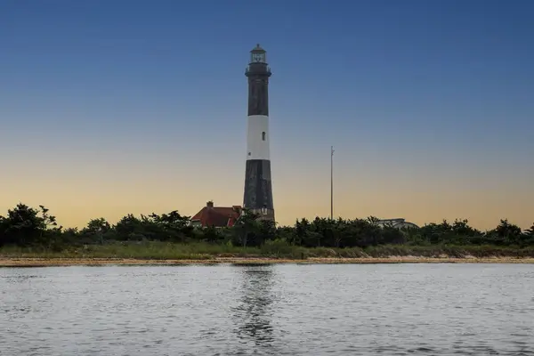 stock image A towering lighthouse stands majestically by the water, surrounded by lush greenery. The serene sky transitions from a warm sunset to a calm blue as it meets the horizon and reflects off the gentle waves.