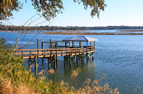 stock image A wooden pier in Bluffton South Carolina extends over a calm body of water, topped with a small roofed structure. In the background, a shoreline and trees give a serene and natural feel to the scene.