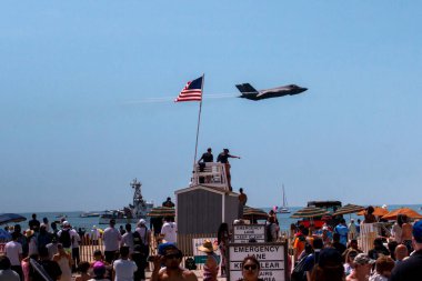 Jones Beach, New York, ABD - 29 Mayıs 2022: Kalabalık bir hava gösterisi sırasında bir F-35 Lighning Steghalth savaş uçağı sahili üzerinde uçuyordu. Büyük bir Amerikan bayrağı bir cankurtaran standında dalgalanırken birkaç kişi ayakta gösteriyi izliyordu..