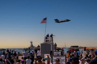 Jones Beach, New York, ABD - 29 Mayıs 2022: Kalabalık bir hava gösterisi sırasında bir F-35 Lighning Steghalth savaş uçağı sahili üzerinde uçuyordu. Büyük bir Amerikan bayrağı bir cankurtaran standında dalgalanırken birkaç kişi ayakta gösteriyi izliyordu..