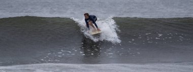 Gilgo Beach, New York, ABD - 30 Ağustos 2023: Bir sörfçü bulutlu bir günde ustalıkla dalgalara binerek okyanusun sularında çeviklik ve kontrol sergilemektedir..